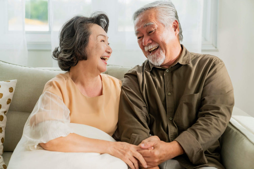 Happy senior Asian couple laughing together while sitting on a sofa at home.